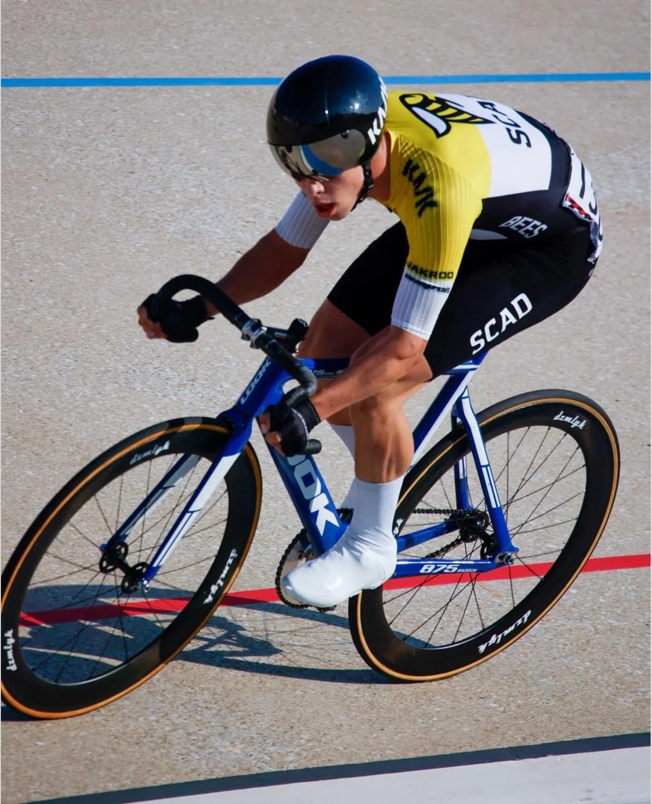 Alejandro racing a track bike for SCAD on a velodrome, in a yellow and black SCAD Bees skinsuit and aero helmet.
